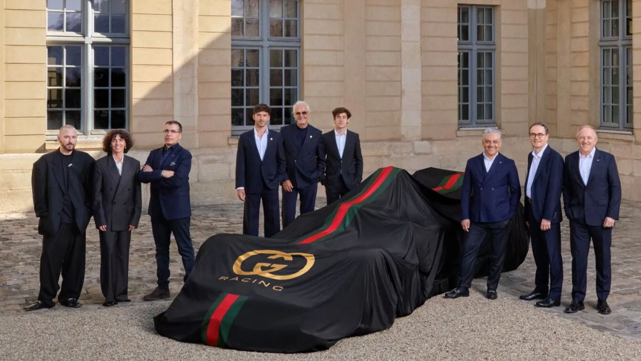 Dieci persone in abito formale posano davanti a un'auto coperta da un telo nero con logo Gucci e strisce verdi e rosse.