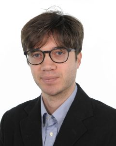 Ritratto di un uomo caucasico con capelli castani, occhi azzurri e occhiali neri, in giacca e camicia.