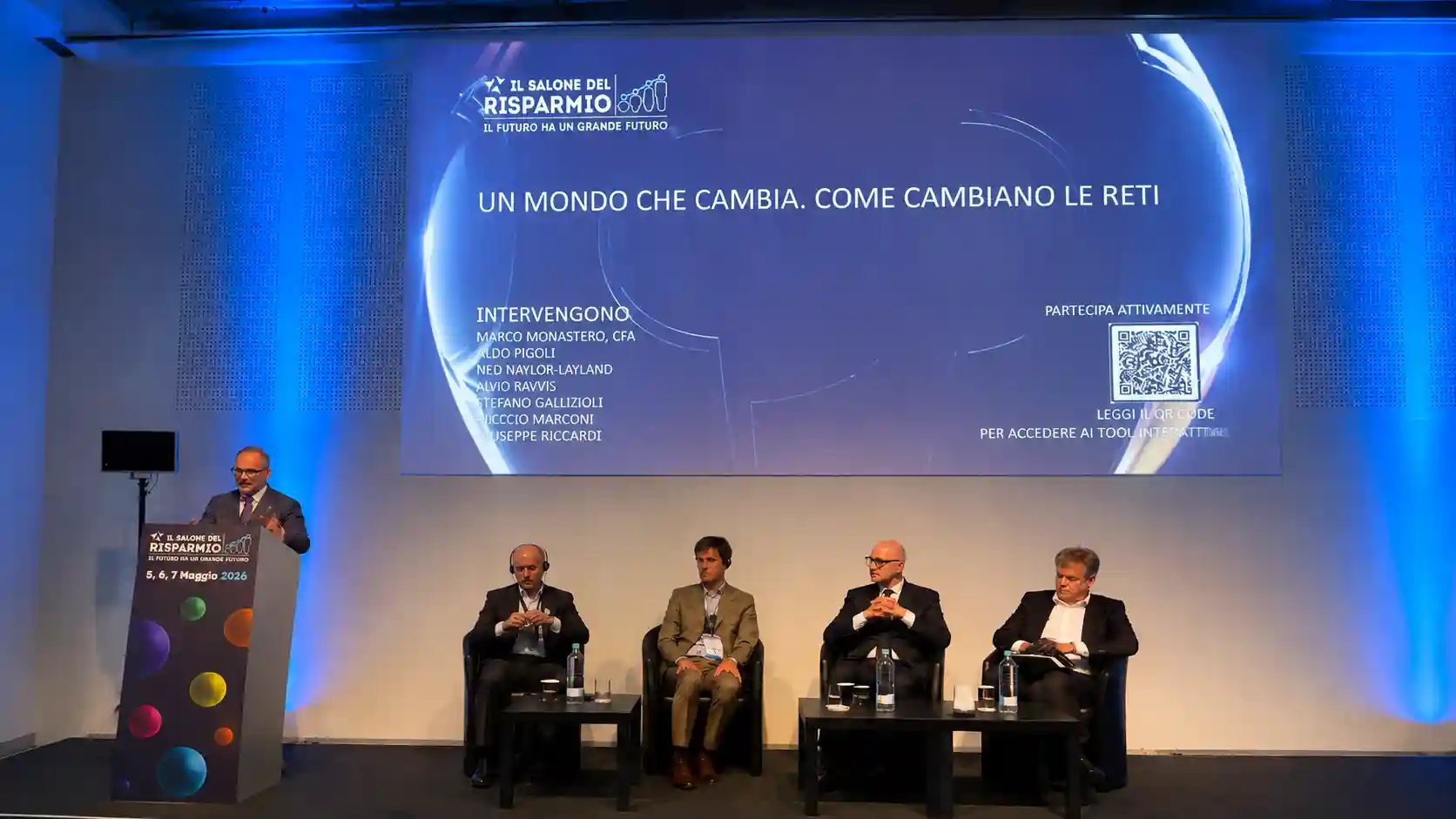 Un uomo parla a un evento con relatori seduti, schermo con titolo "Un mondo che cambia".