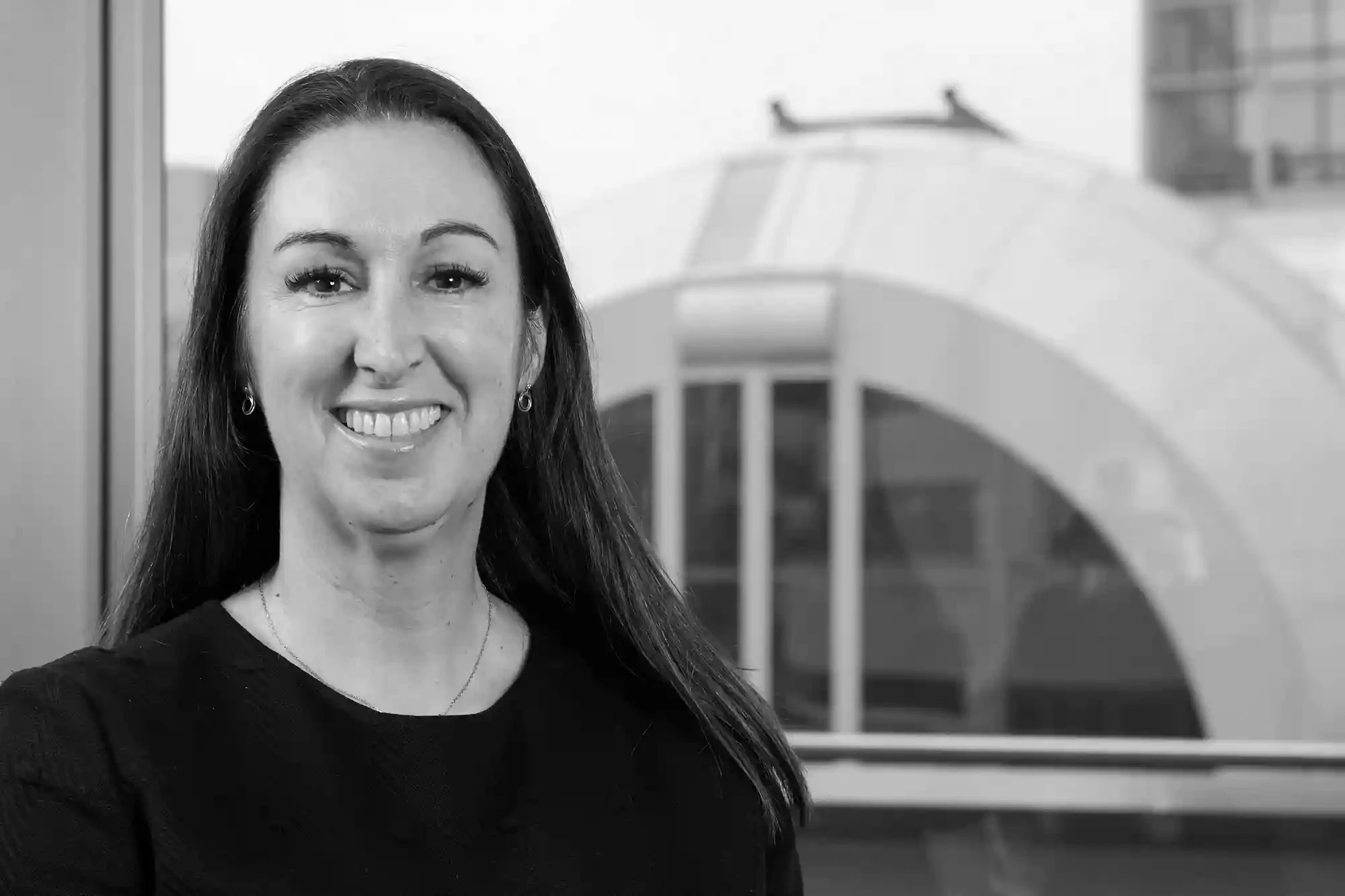 Ritratto in bianco e nero di una donna sorridente con lunghi capelli scuri, edificio moderno sullo sfondo.