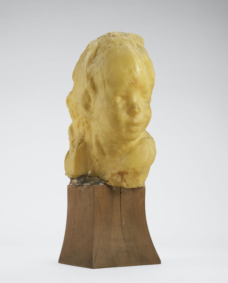 Scultura di cera gialla raffigurante il busto di un bambino, su base di legno marrone.