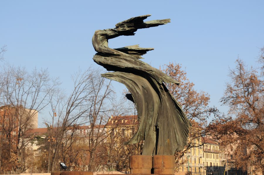Scultura in bronzo stilizzata di figura umana che si protende verso l'alto, con edifici e alberi sullo sfondo.