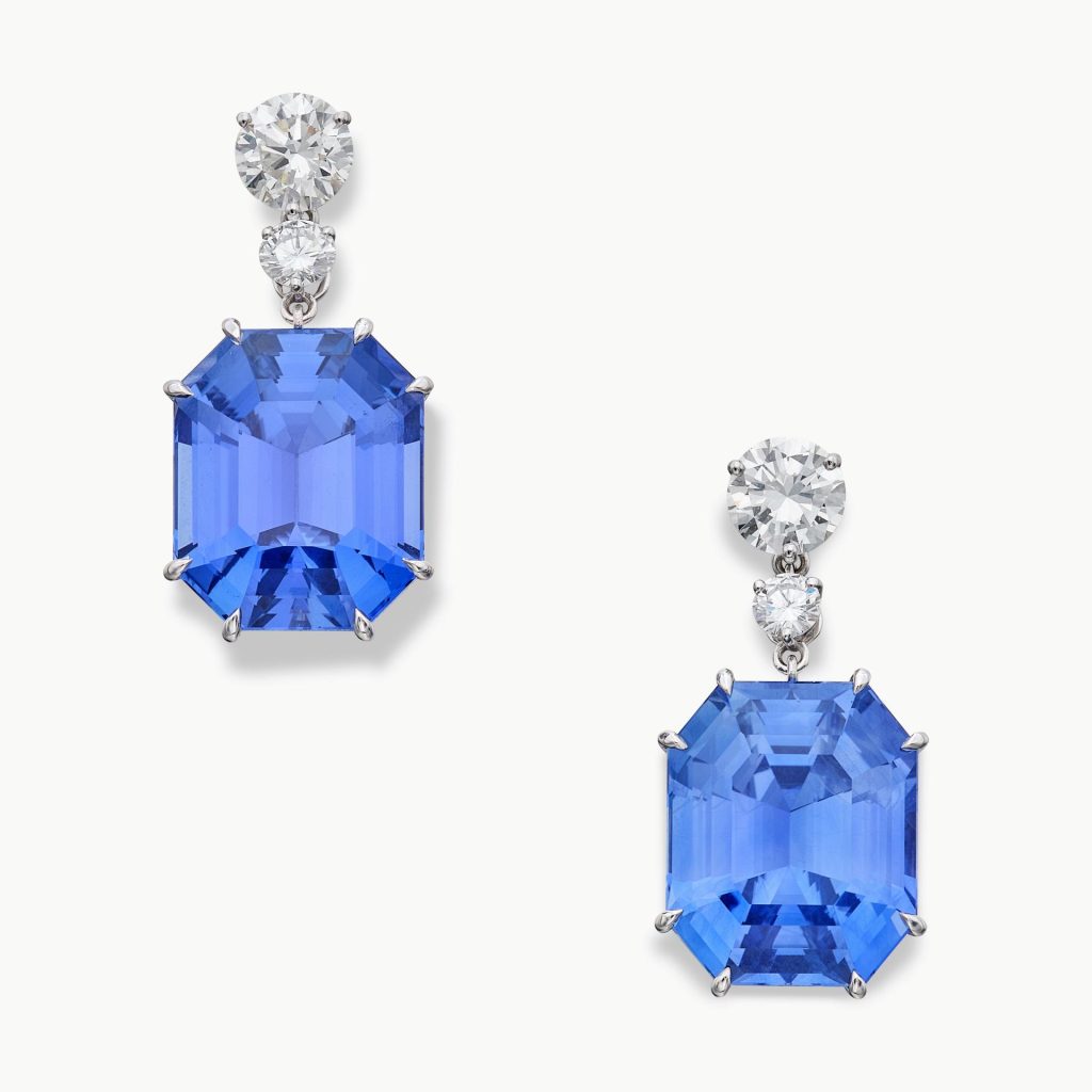Due orecchini pendenti con zaffiri blu taglio smeraldo e diamanti su sfondo bianco.