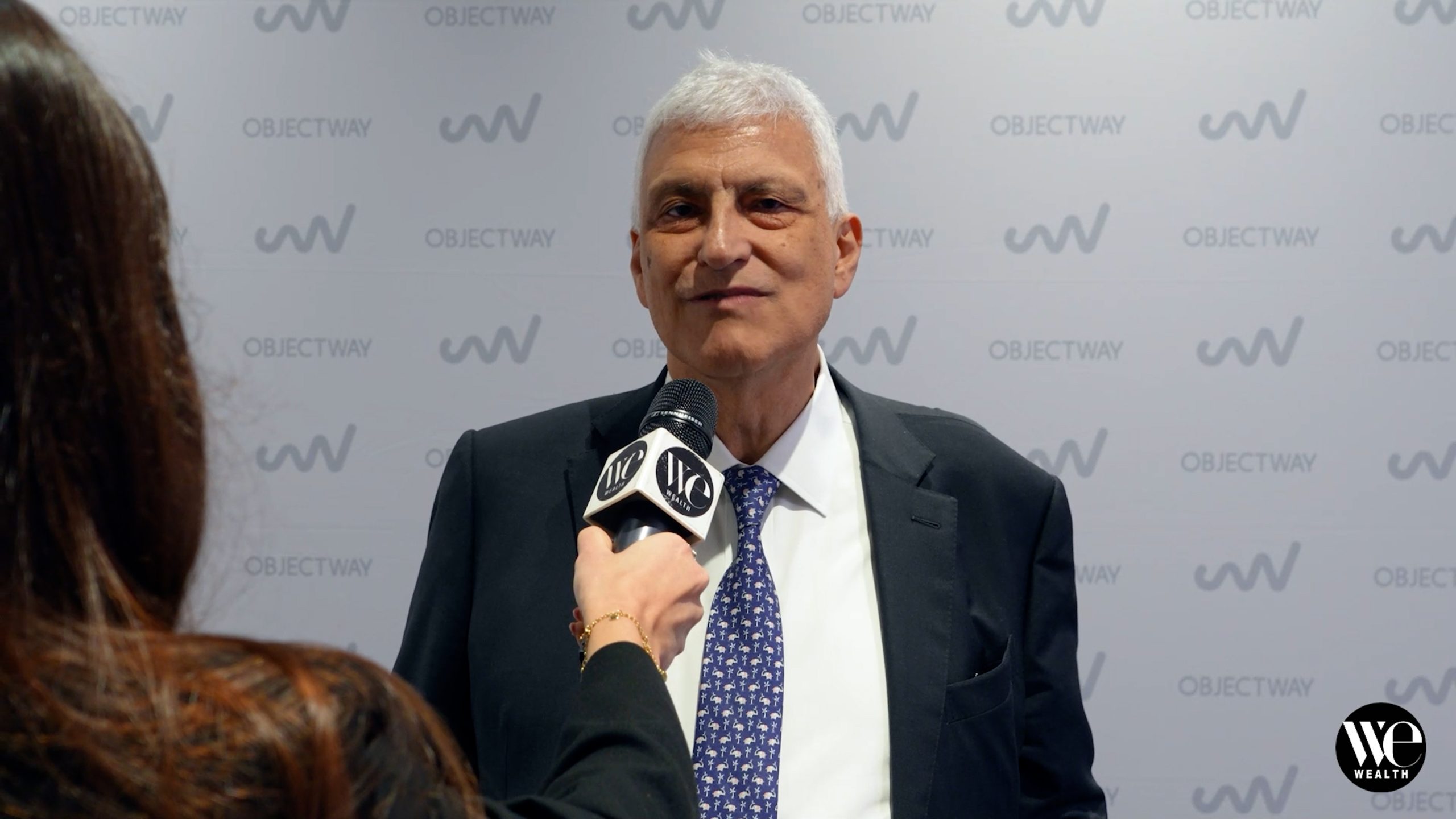 Luigi Marciano di Objectway intervistato con microfono davanti a sfondo con logo "Objectway" e "WE Wealth". Indossa giacca scura e cravatta blu.