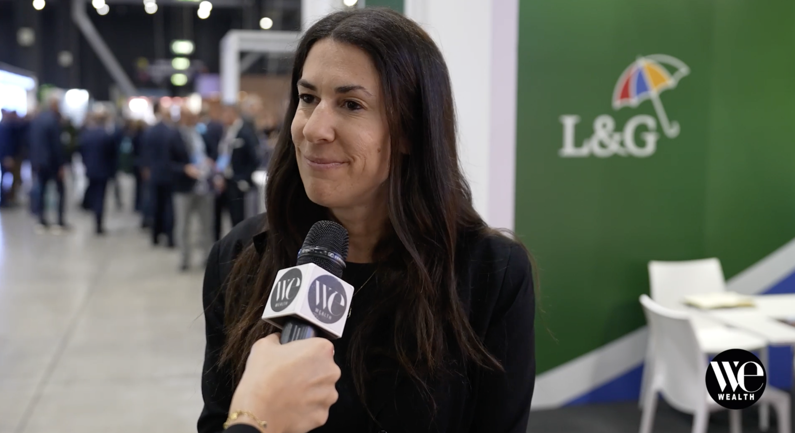 Elena Baccani di L&G intervistata con microfono "WE Wealth" davanti a stand verde con logo ombrello colorato.