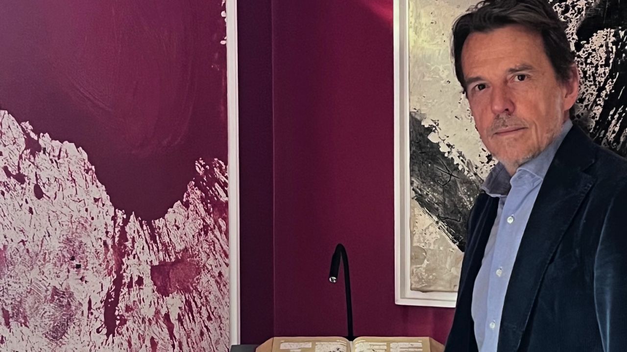 Un uomo con una giacca scura è in piedi in una stanza dalle pareti rosso scuro, accanto a una scrivania con un libro aperto sulla collezione "Come si valuta" e una lampada. Un'opera d'arte astratta con motivi bianchi e rossi è appesa alla parete dietro di lui.