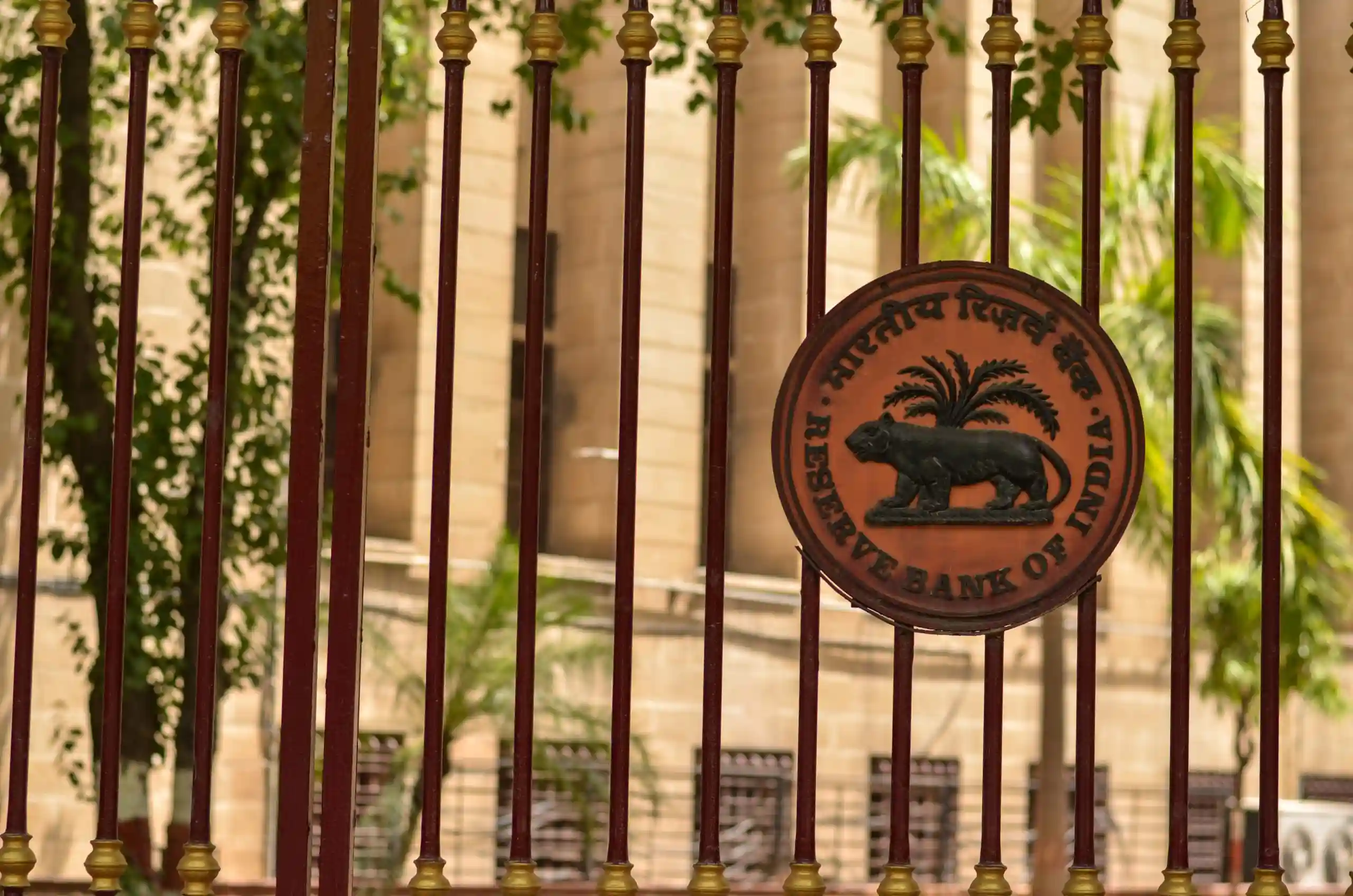 Cancello in ferro battuto marrone con stemma rotondo della Reserve Bank of India, vegetazione sullo sfondo.