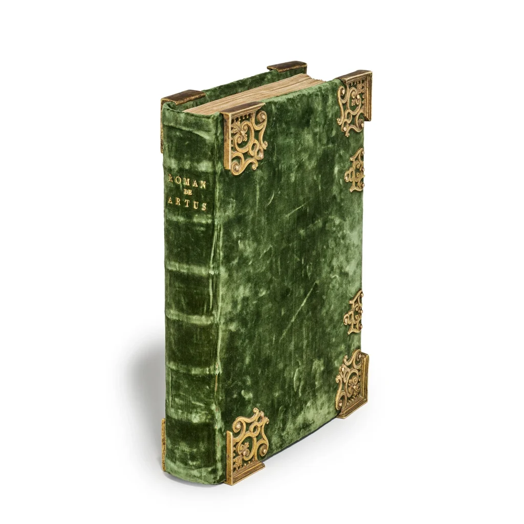 Libro antico "Roman de Artus" con copertina in velluto verde e decorazioni angolari dorate su sfondo bianco.