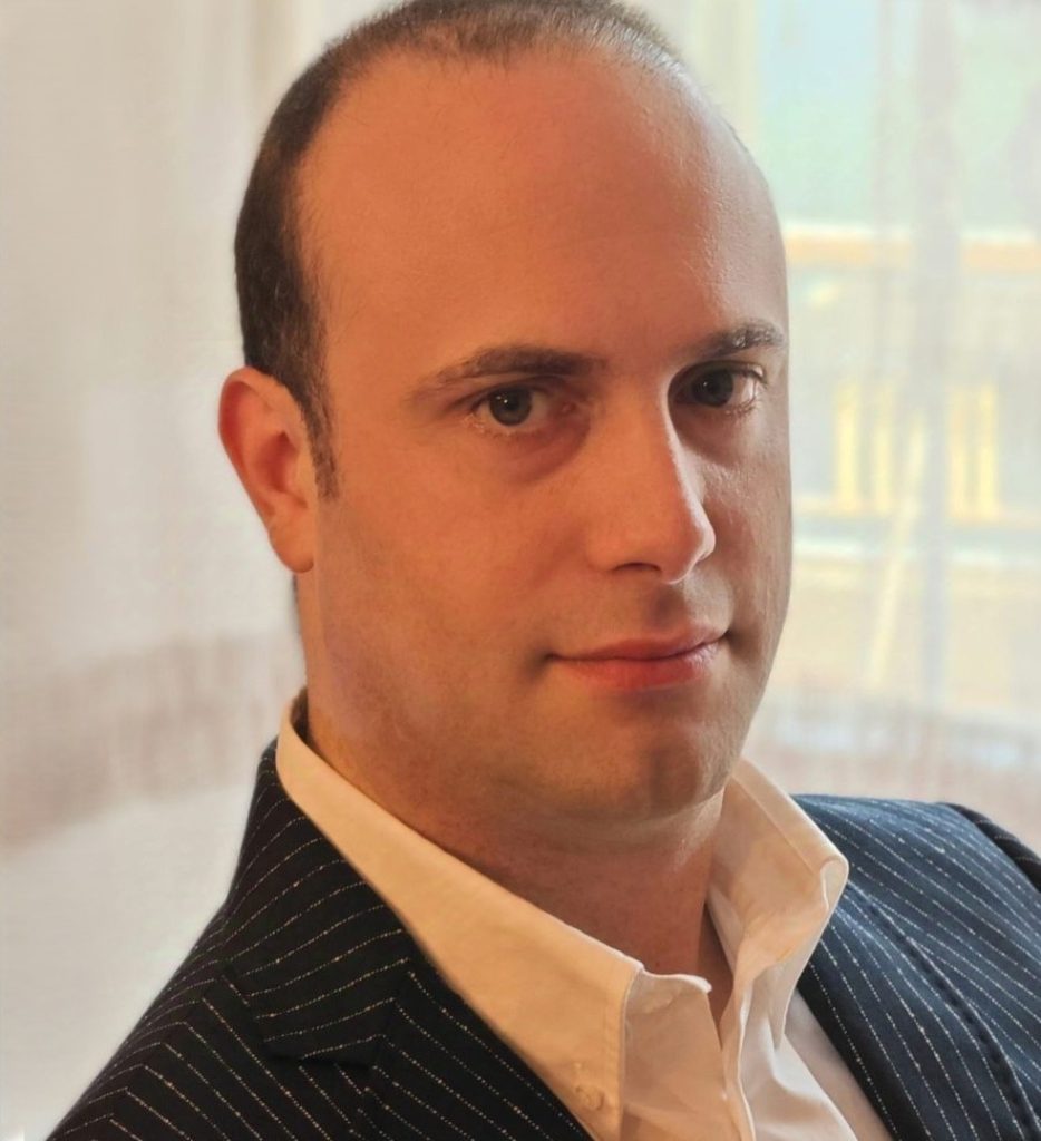 Ritratto di uomo calvo con camicia bianca e giacca gessata, sguardo diretto e sfondo luminoso.