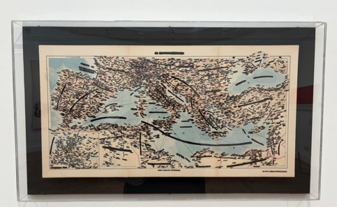 Mappa geografica con aree cancellate da segni neri, incorniciata in una teca trasparente con sfondo nero.