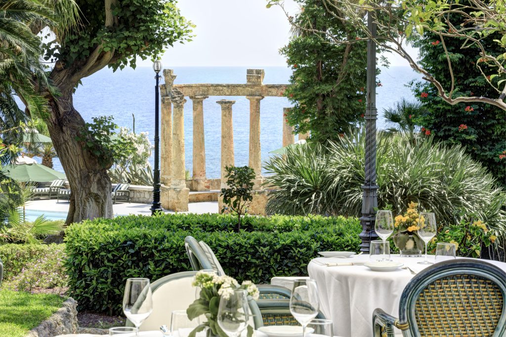 Tavolo apparecchiato con vista sul mare, colonne antiche e vegetazione lussureggiante.