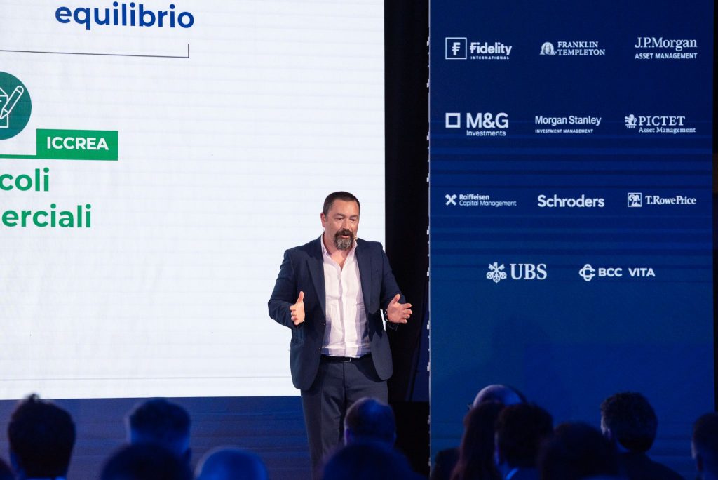 Uomo con barba parla su un palco, con loghi aziendali sullo sfondo.