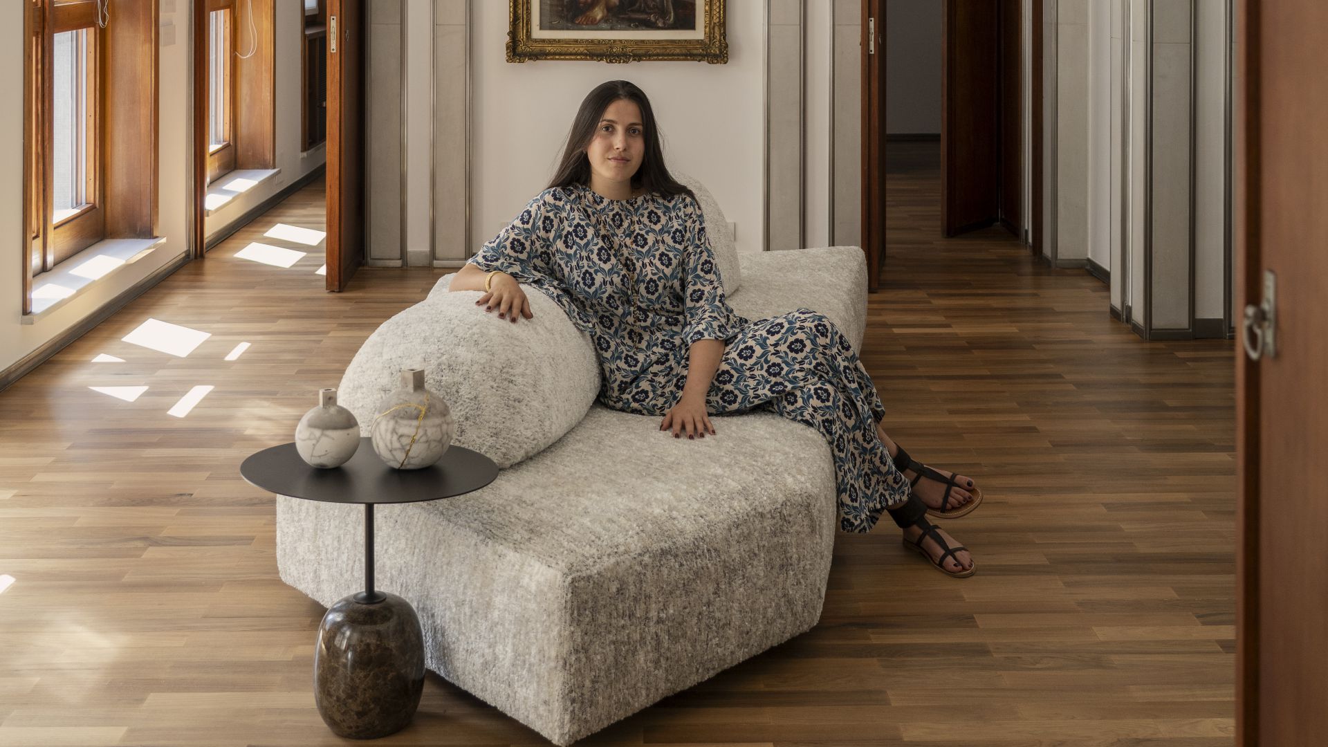 Donna con abito floreale seduta su divano bouclé, tavolino con vasi, parquet e luce solare.
