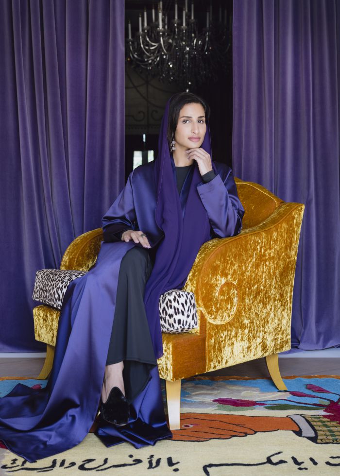Donna con velo viola, abito blu, seduta su poltrona dorata, con tende viola e tappeto colorato.