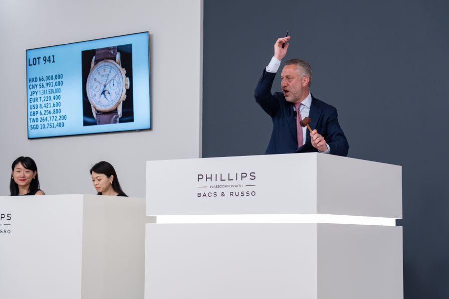 Asta di orologi: banditore con martelletto, schermo con prezzi in valute diverse e due donne.