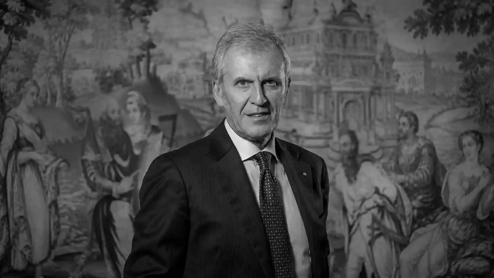 Uomo in abito scuro con cravatta, sorride davanti a un arazzo con figure storiche.