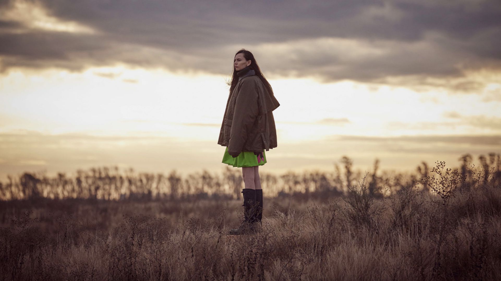 Una persona con un abito verde, una giacca marrone e stivali neri è in piedi in un campo erboso al tramonto, evocando lo stile cinematografico di Miu Miu, tra un cielo nuvoloso e alberi in lontananza sullo sfondo.