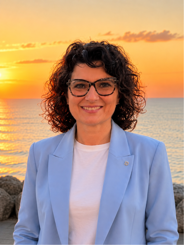 Donna con occhiali sorride con un blazer azzurro, tramonto sul mare sullo sfondo.