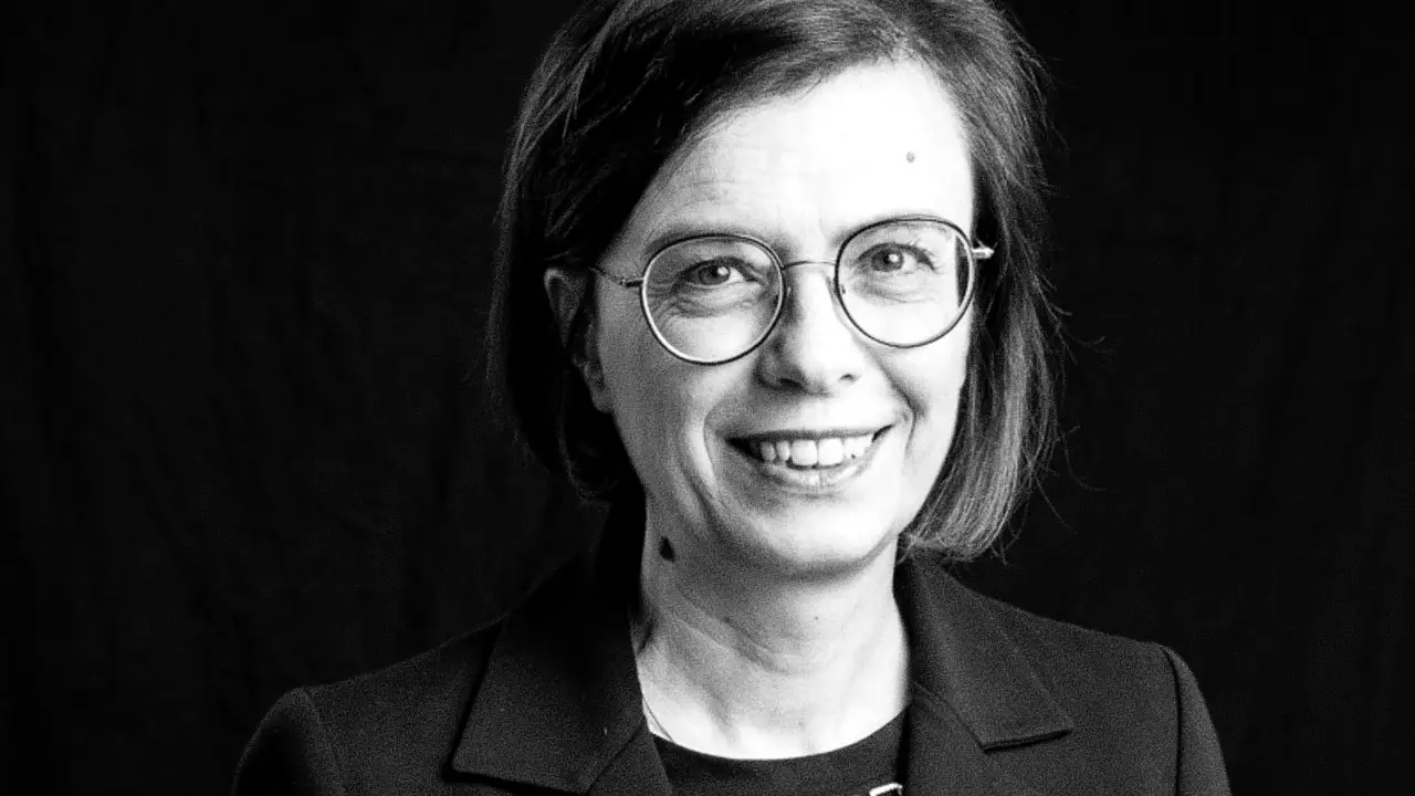 Donna con occhiali rotondi sorride, indossa una giacca scura.