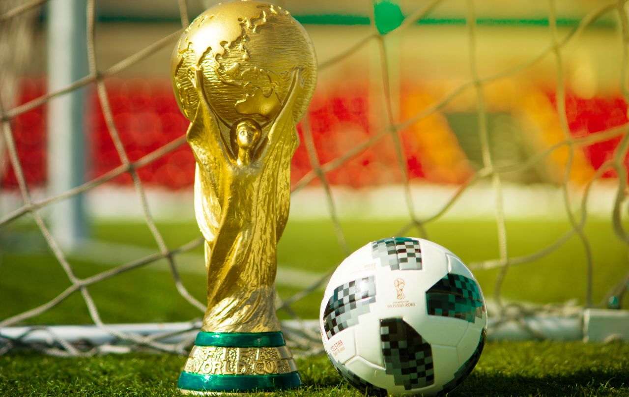 Coppa del Mondo dorata e pallone da calcio su erba verde, con rete da calcio e tribune sfocate sullo sfondo.