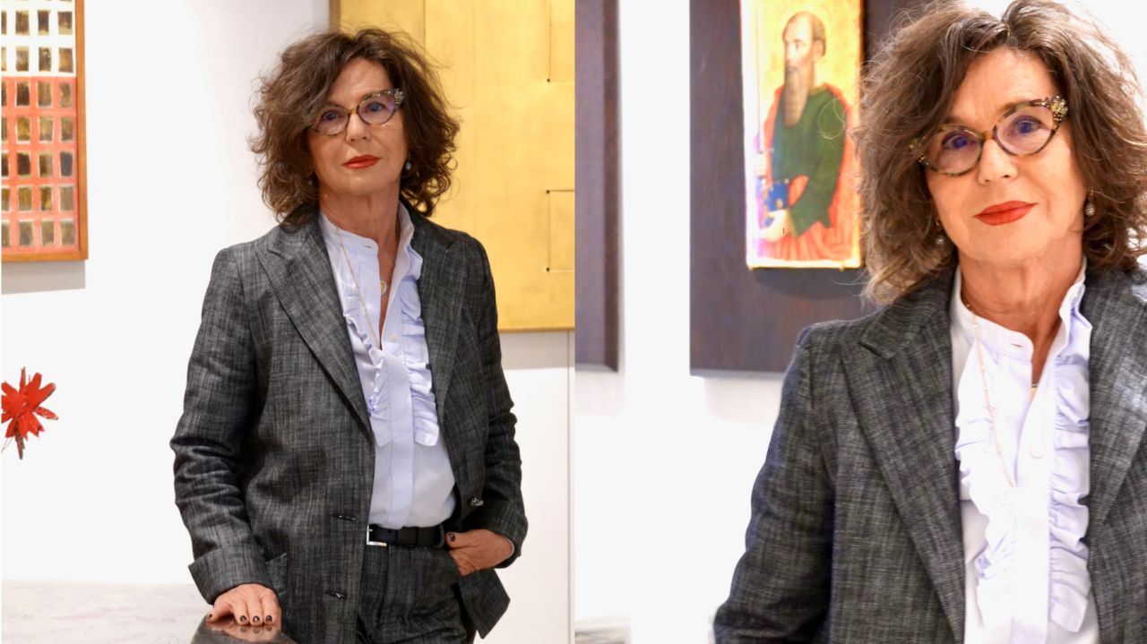 Donna con capelli mossi, occhiali, tailleur grigio, camicia con volant, in una galleria d'arte.