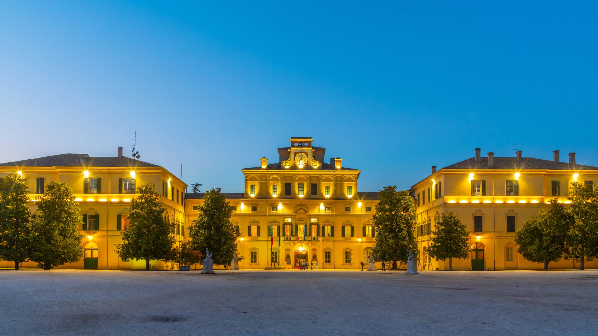 Palazzo giallo illuminato con un grande orologio, alberi verdi e cielo blu crepuscolare.