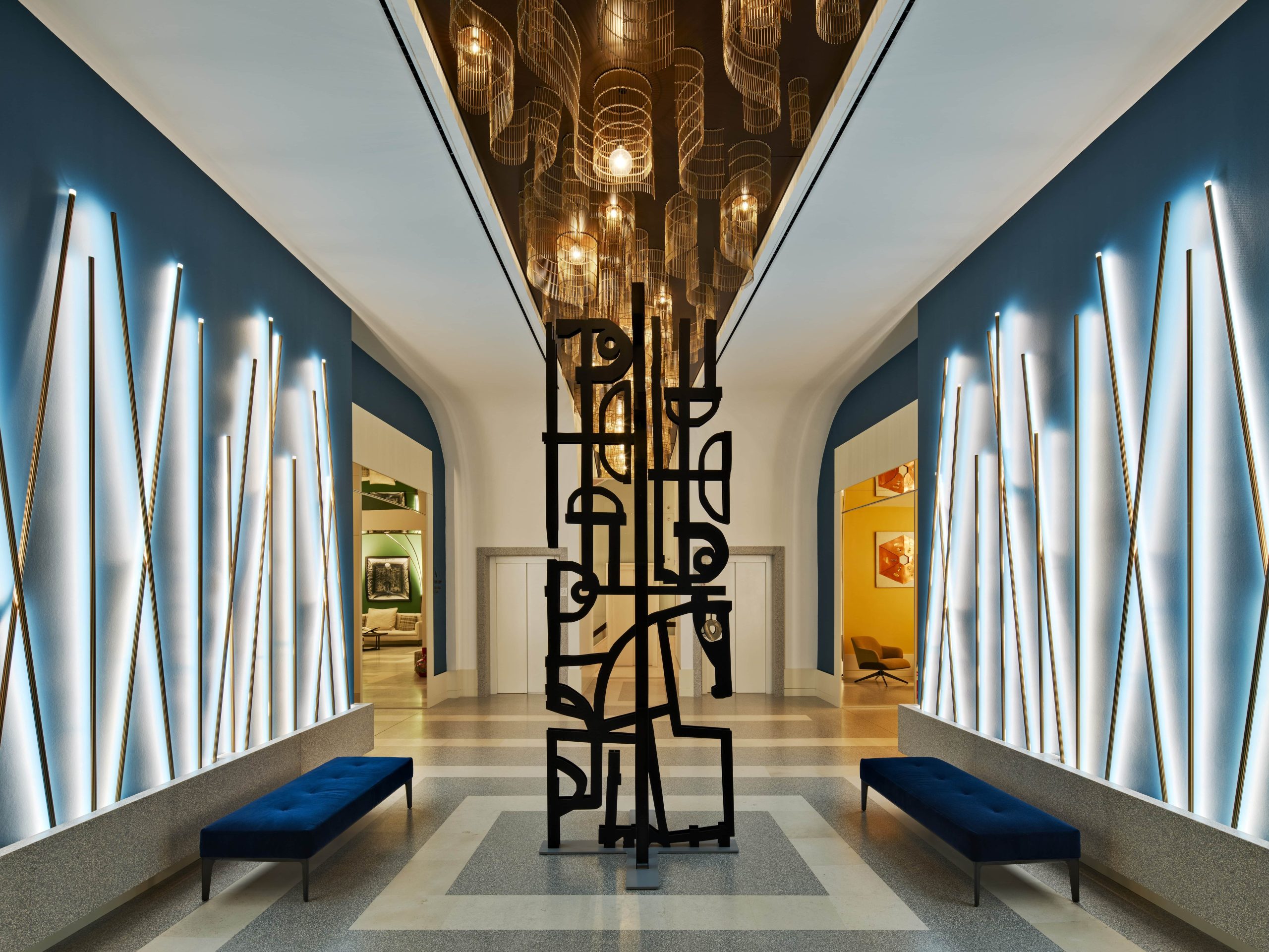 Corridoio moderno con scultura astratta, panchine blu e illuminazione a parete.
