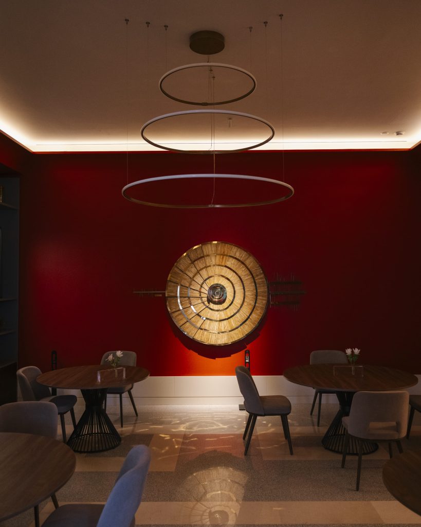 Tavoli rotondi, sedie grigie, decorazione murale circolare in legno e metallo, luci a sospensione.