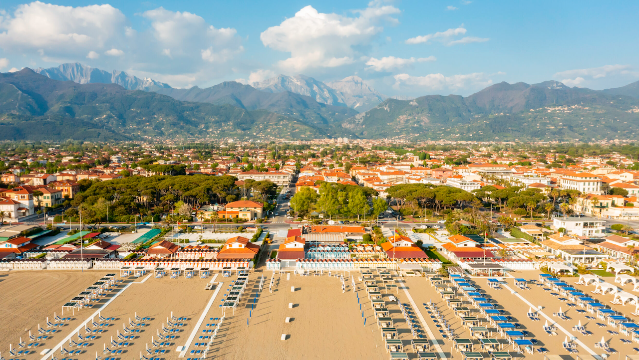 Spiaggia con file di lettini e ombrelloni, città e montagne sullo sfondo.