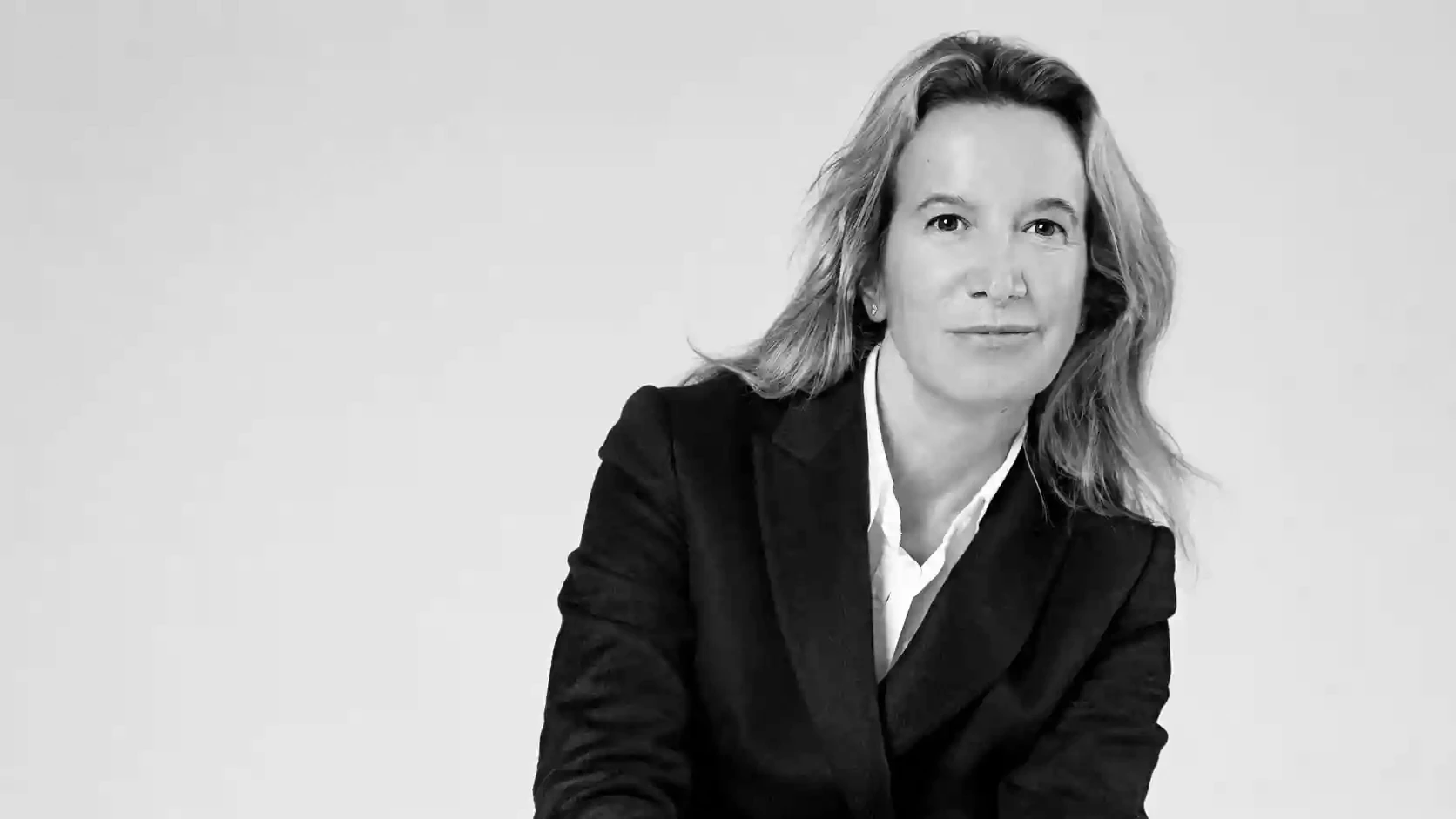 Donna con capelli biondi lunghi, indossa un tailleur nero e camicia bianca, guarda verso la telecamera.