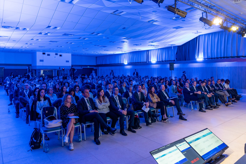 Pubblico seduto in una sala conferenze illuminata di blu, con schermi in primo piano.