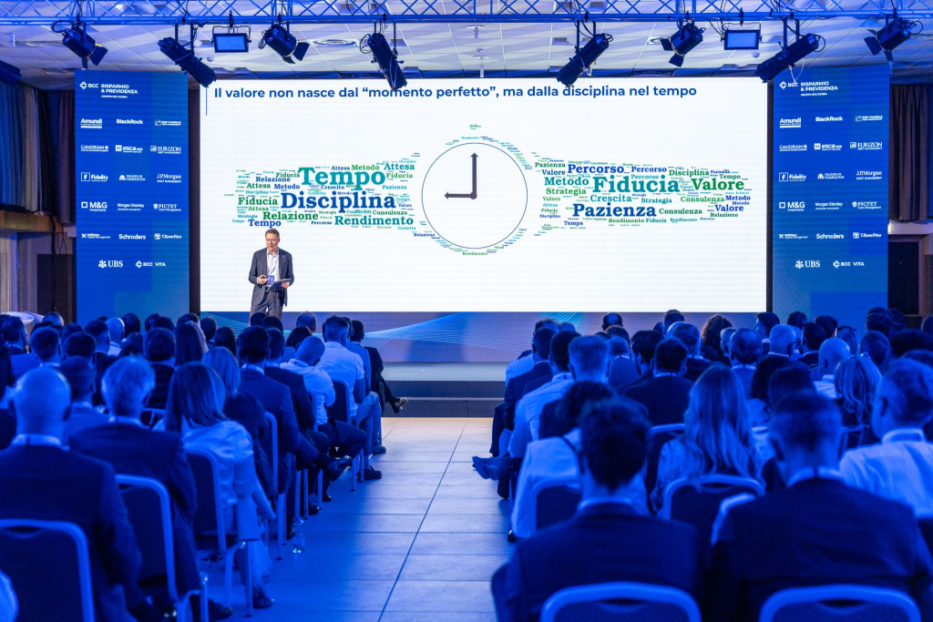 Uomo parla a un pubblico seduto davanti a uno schermo con una nuvola di parole a forma di orologio.