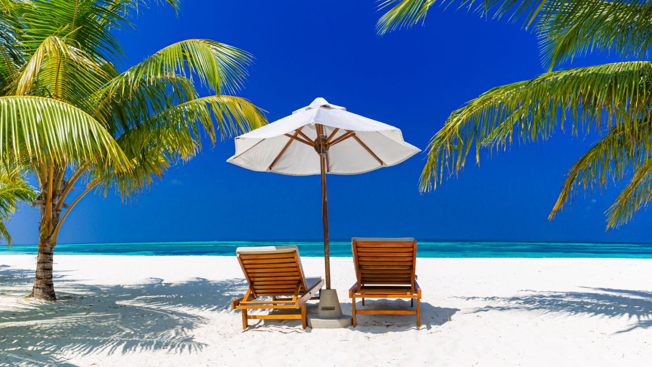 Due lettini prendisole e un ombrellone bianco su una spiaggia tropicale con palme.