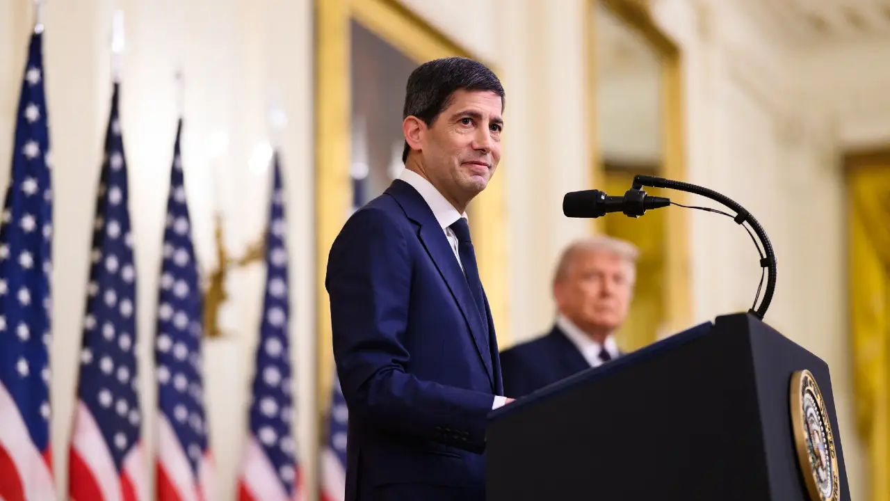 Uomo in abito blu parla al microfono, con Donald Trump sullo sfondo e bandiere americane.