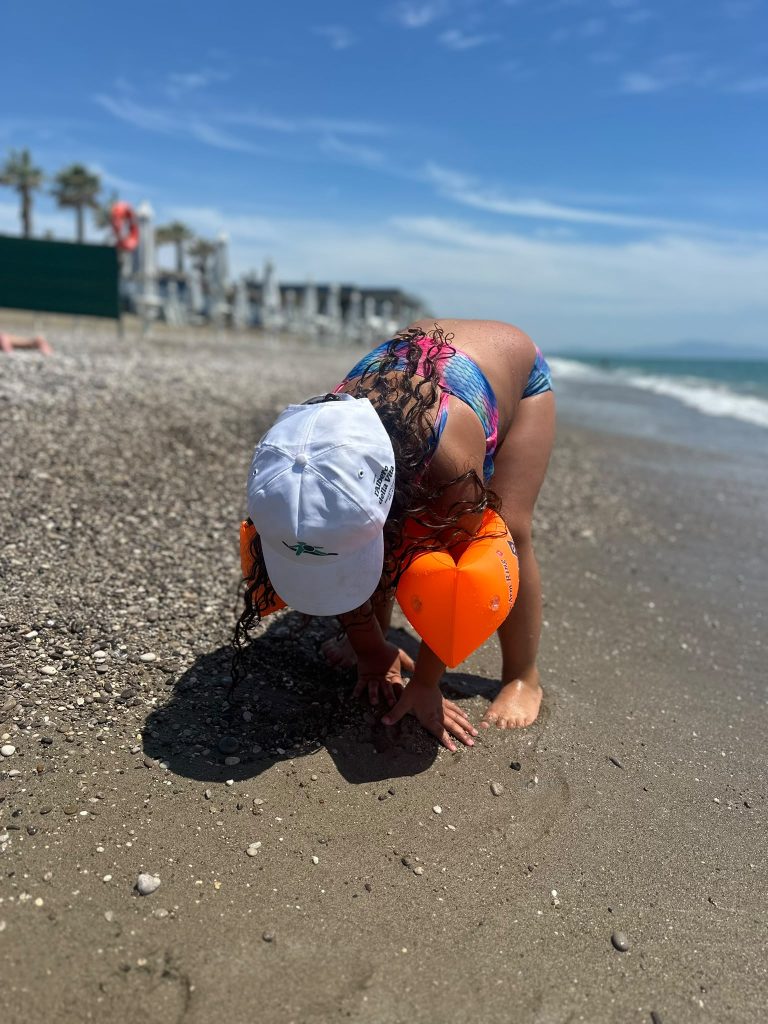 Bambina con braccioli arancioni a forma di cuore raccoglie sassolini sulla spiaggia.