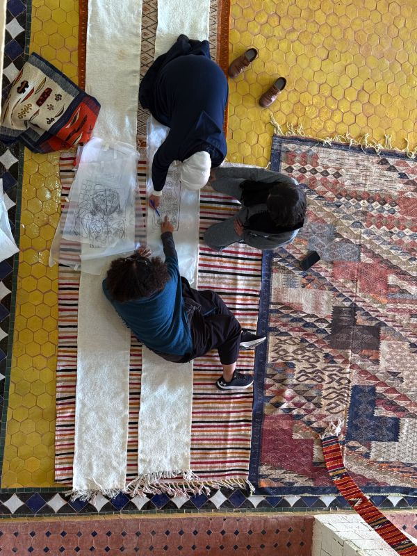 Due persone lavorano su un disegno su tessuto, circondate da tappeti colorati e piastrelle.