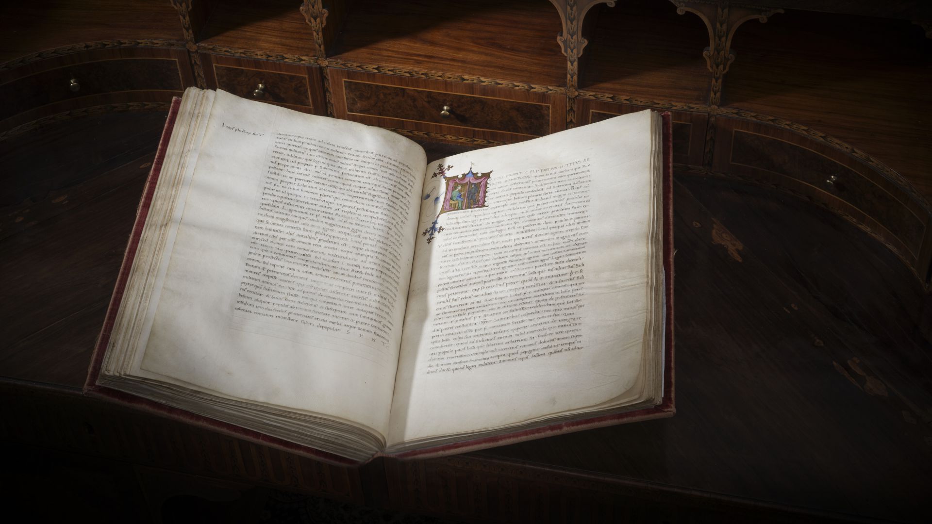 Antico libro aperto con testo manoscritto e un'illustrazione colorata su una scrivania di legno intarsiato.