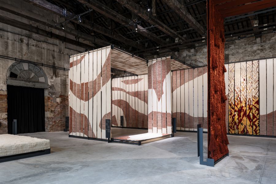 Installazione con pannelli tessili a motivi astratti e un grande arazzo rosso.