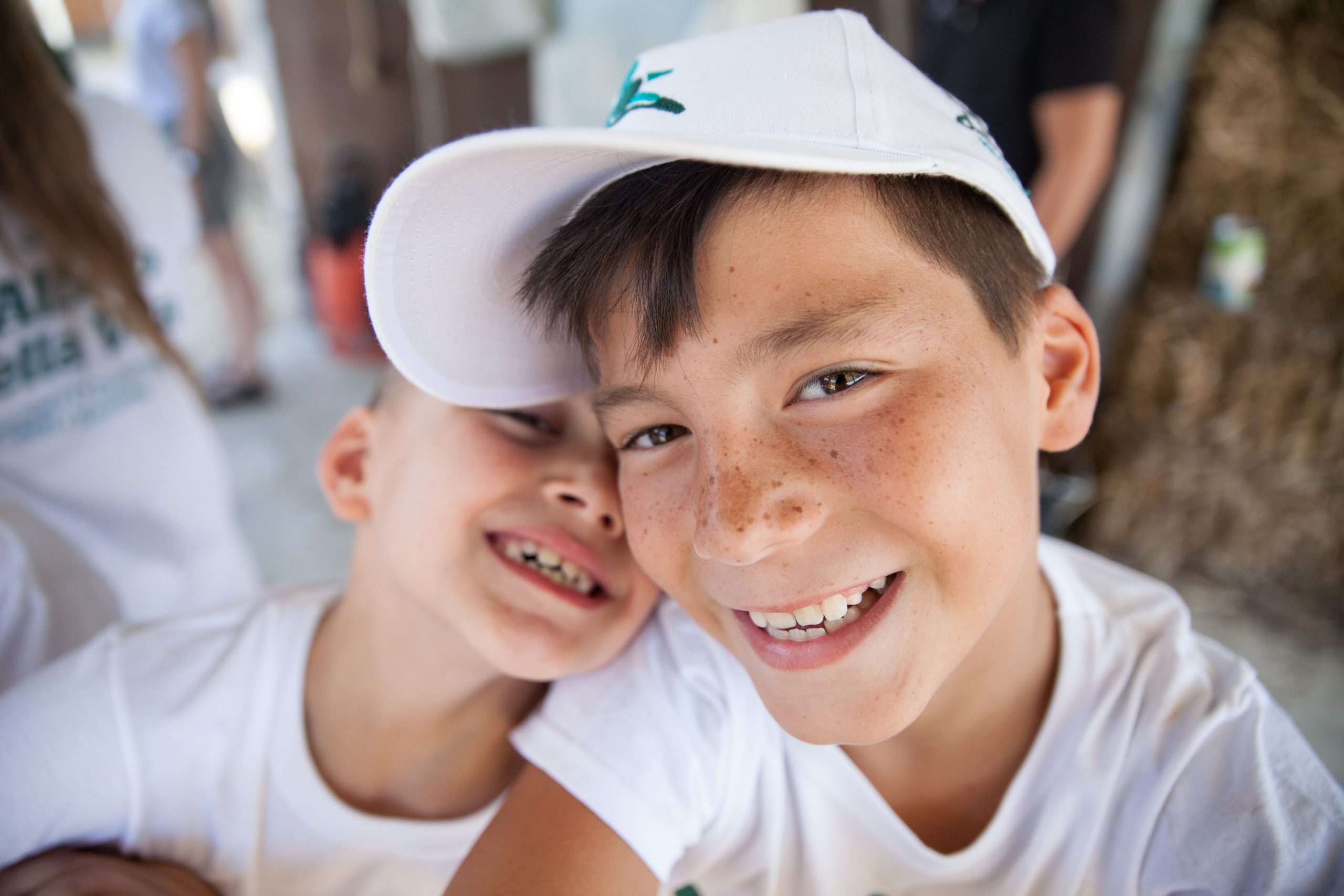 Due bambini sorridono vicini, uno con un cappellino bianco, entrambi con magliette bianche.