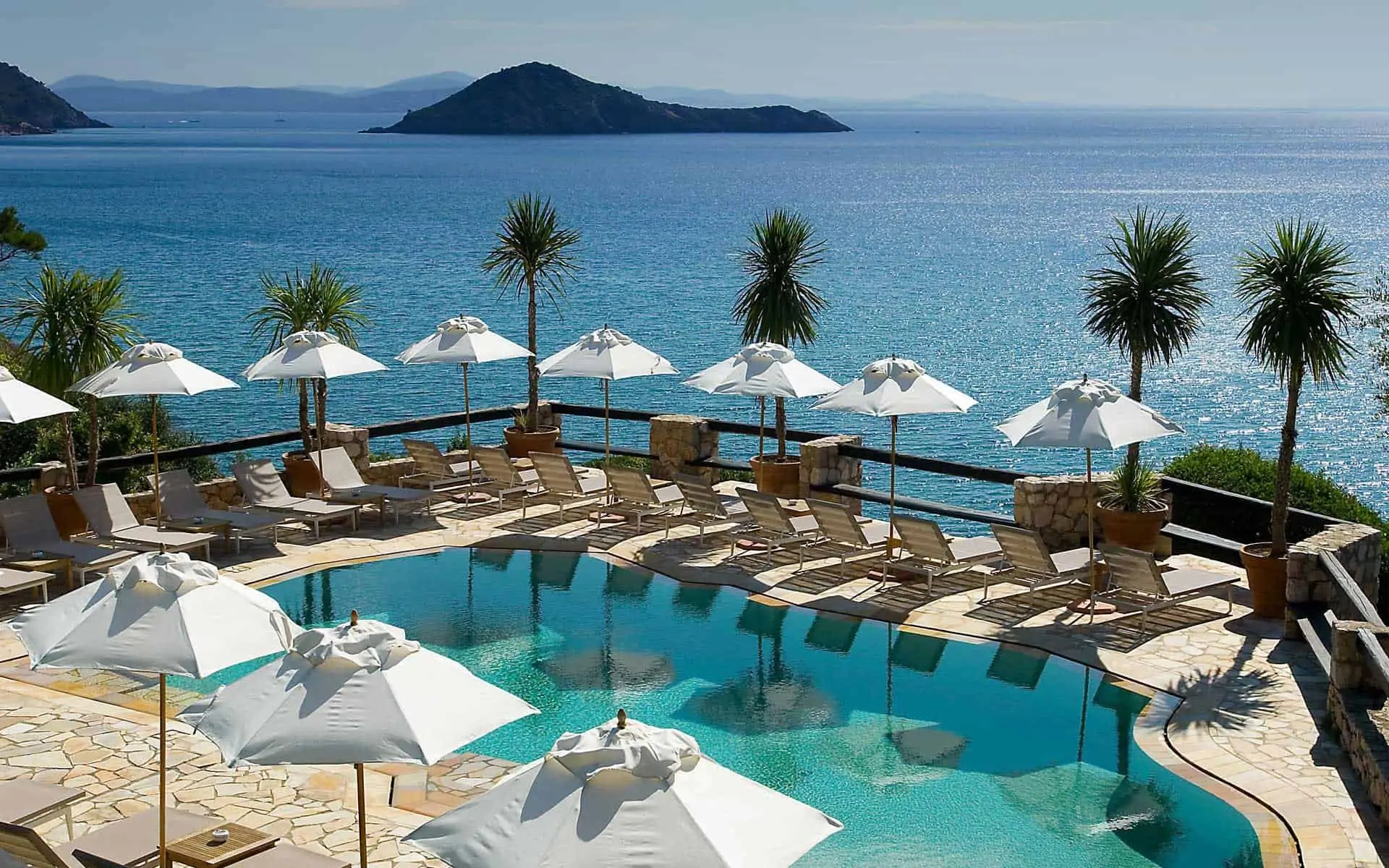 Piscina con lettini e ombrelloni bianchi, palme e mare blu sullo sfondo.