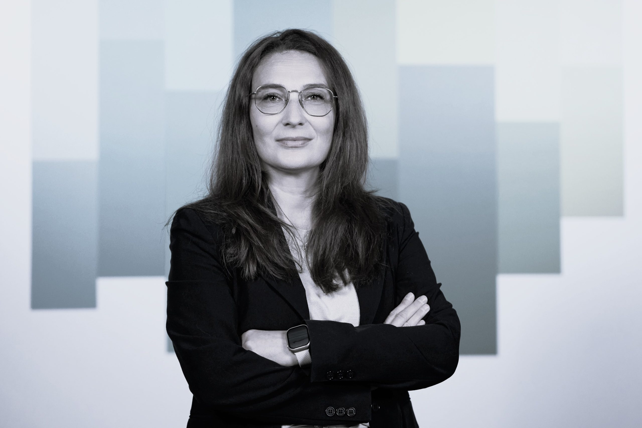 Donna con occhiali e giacca nera, braccia conserte, sfondo geometrico.