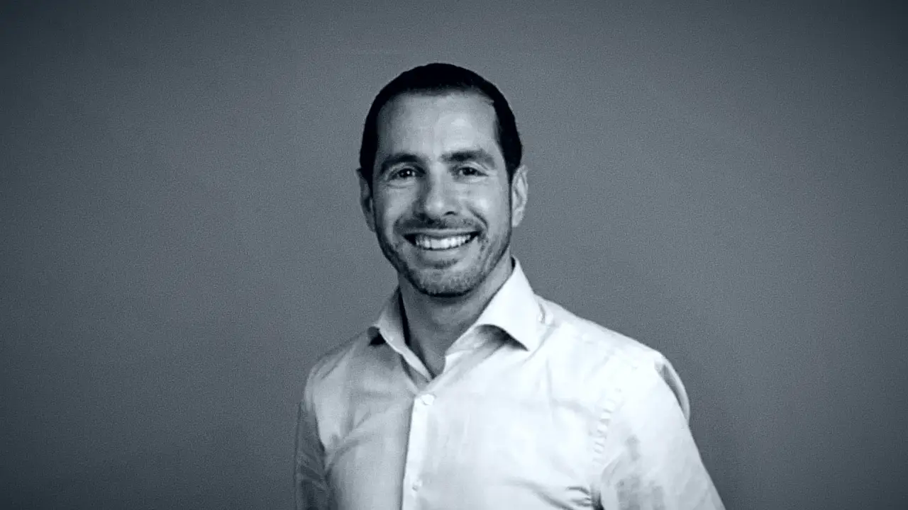 Uomo sorridente in camicia bianca, con sfondo grigio.