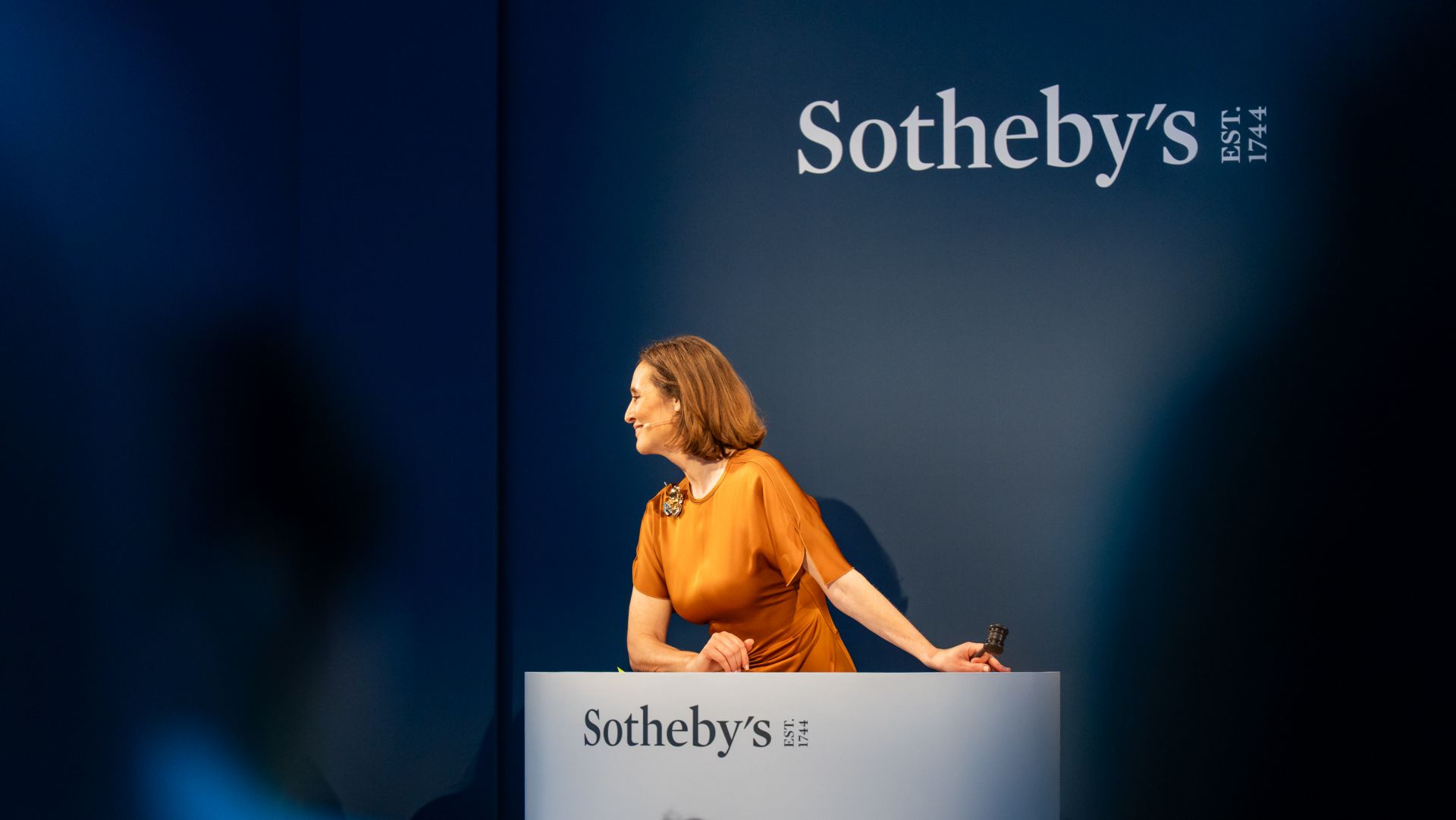 Donna con abito arancione sorride, appoggiata a un podio con logo Sotheby's.