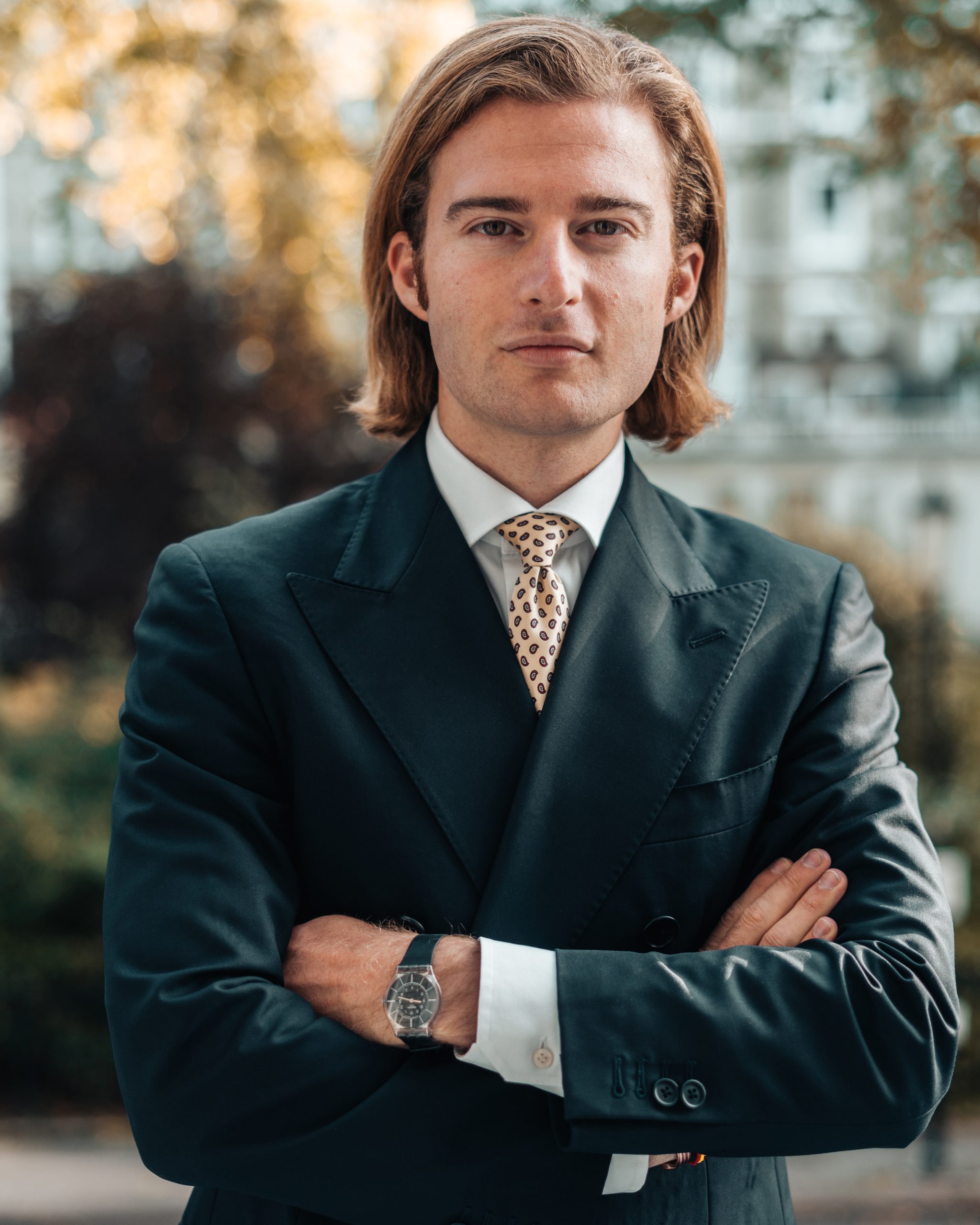 Uomo con capelli biondi lunghi, abito scuro, camicia bianca e cravatta con fantasia.