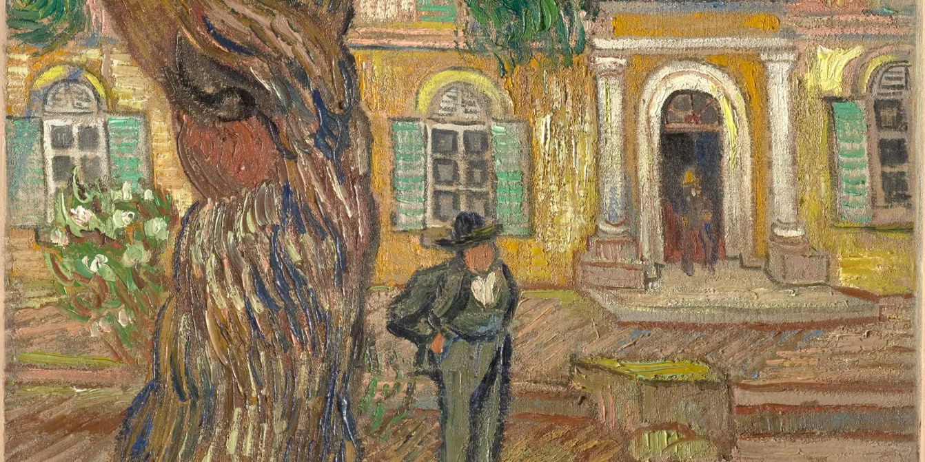 Uomo con cappello in piedi davanti a un edificio giallo con finestre verdi e un grande albero.