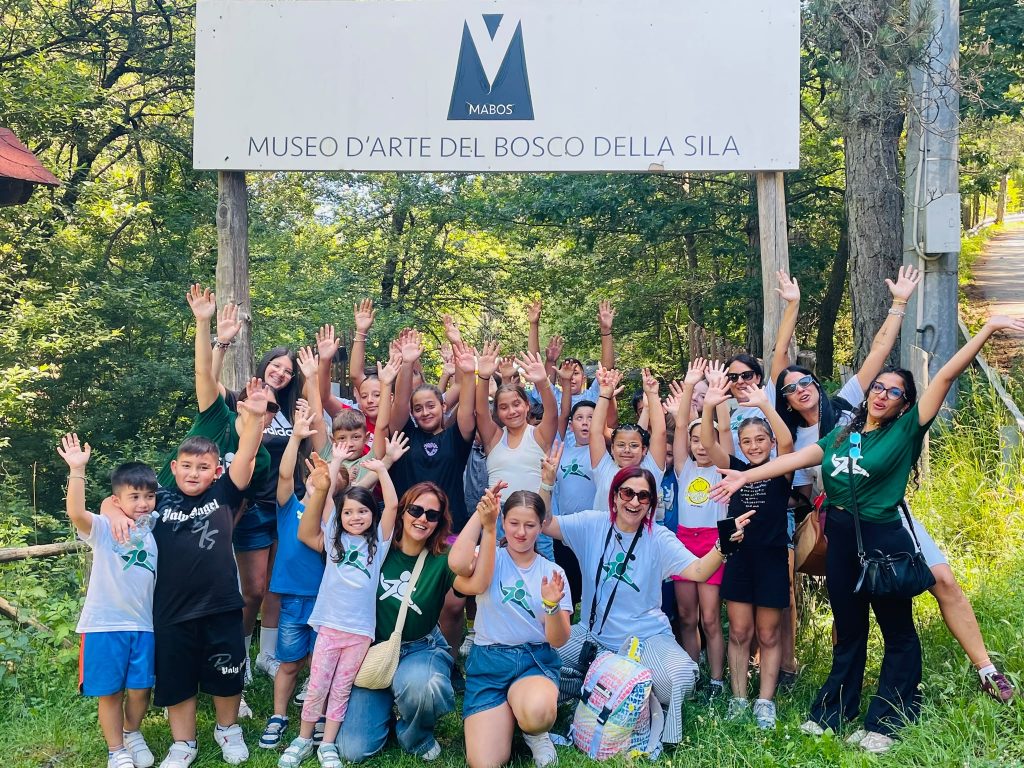 Gruppo di bambini e adulti sorridenti con le mani alzate davanti a un cartello del museo.