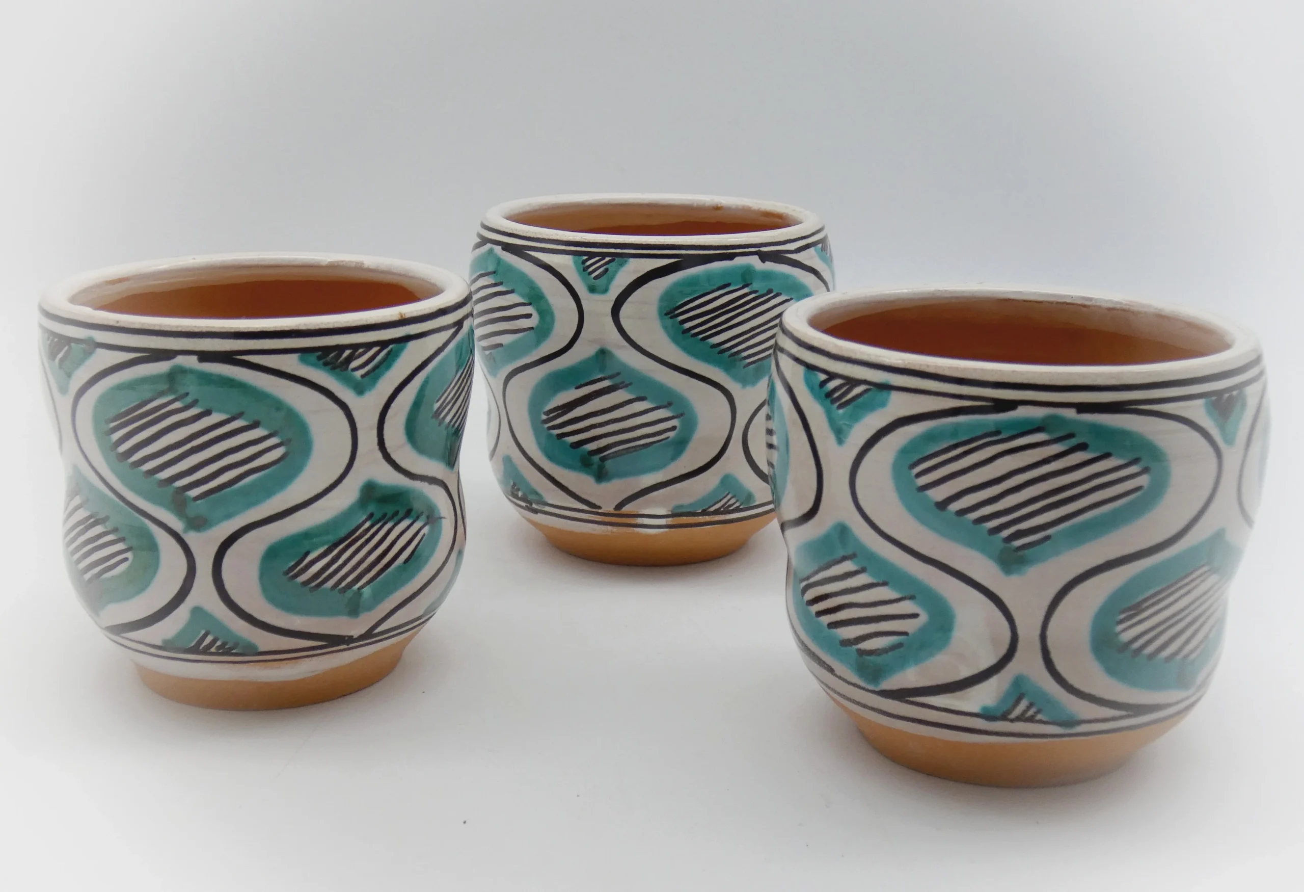 Tre tazze in ceramica con decorazioni geometriche turchesi e nere su sfondo bianco.