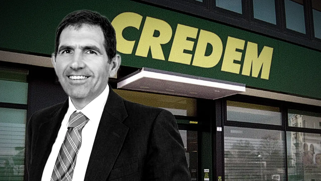Uomo in abito scuro sorride davanti all'insegna verde con scritte gialle "CREDEM".