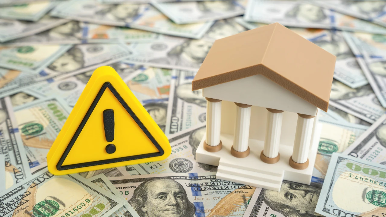 Segnale di pericolo giallo accanto a un modello di edificio bancario su banconote da cento dollari.
