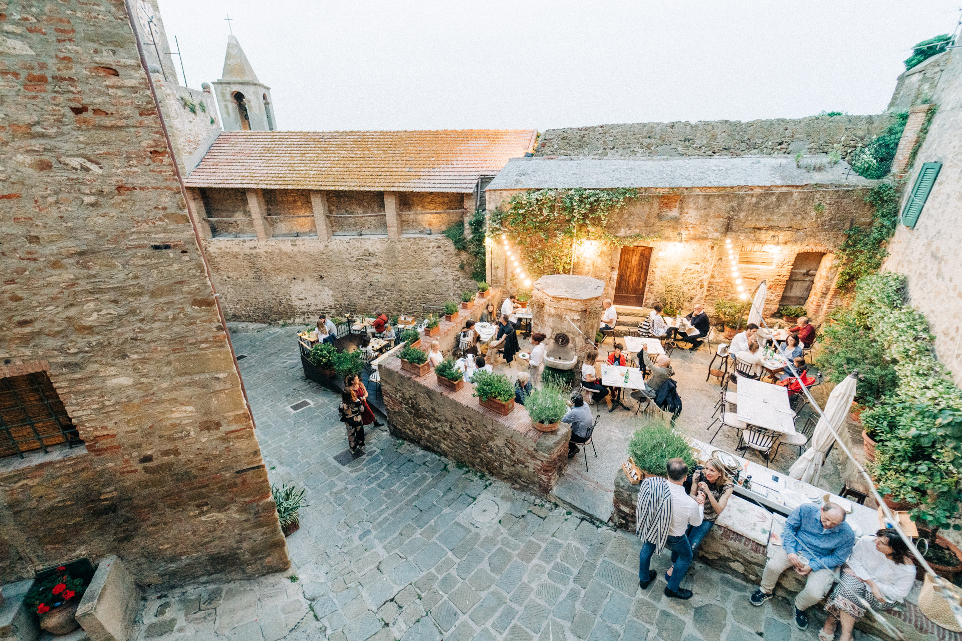 Persone sedute a tavoli all'aperto in un cortile di pietra con luci decorative.